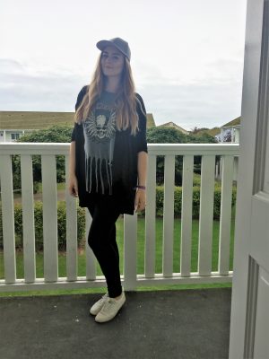 me posing outside the flat with a grey cap, grey skull top, black jeans, black cardigan and white trainers